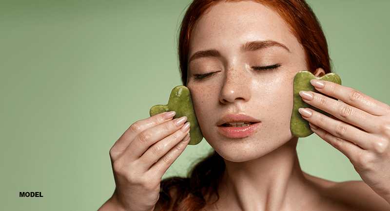 woman performing gua sha on her face using two jade stone tools